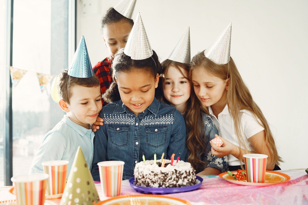Illustrative stock photo of five children in party hats gathered around a birthday cake beside a bright window