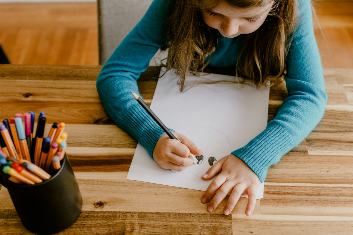 A child draws at a wooden table beside a cup of colored pencils